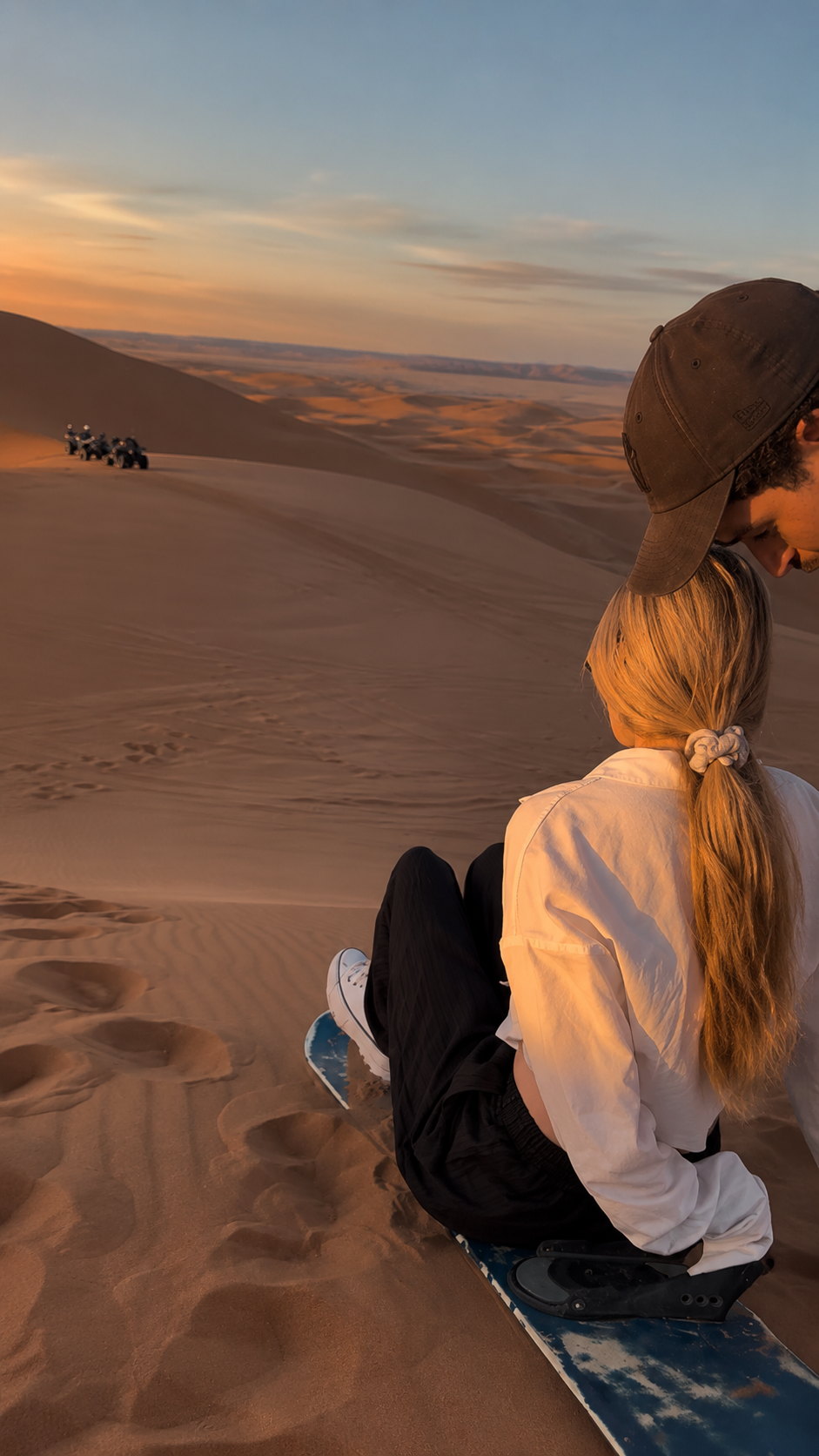 Dos viajeros sentados en una tabla de sandboard viendo la puesta de sol sobre las dunas